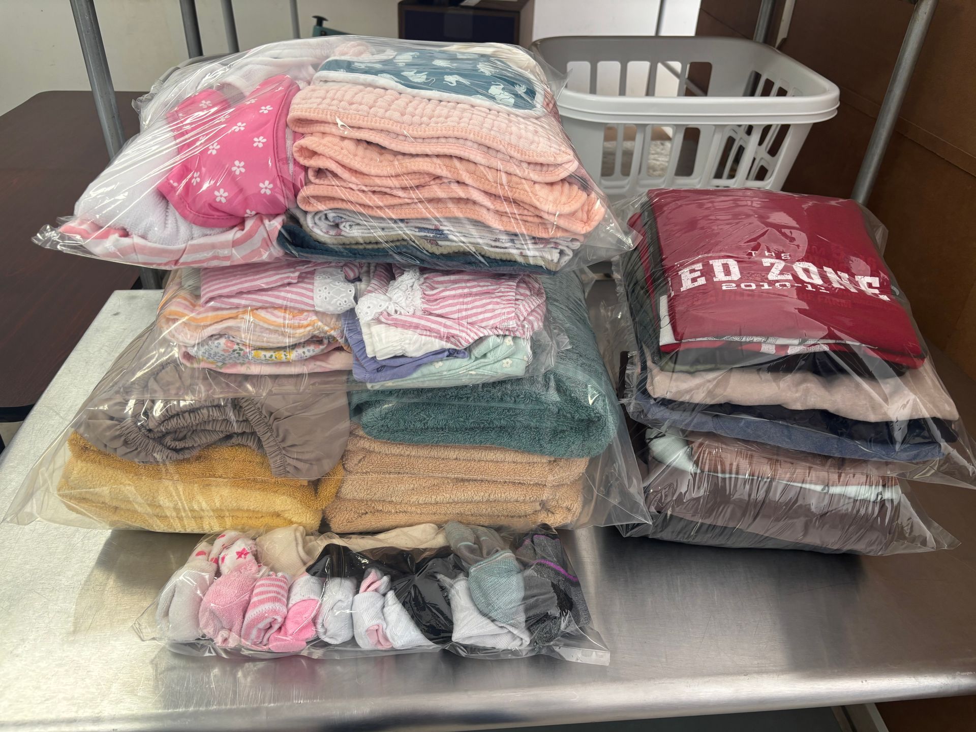 Several clear plastic bags stacked with folded clothing on a metal table. A laundry basket is also present.