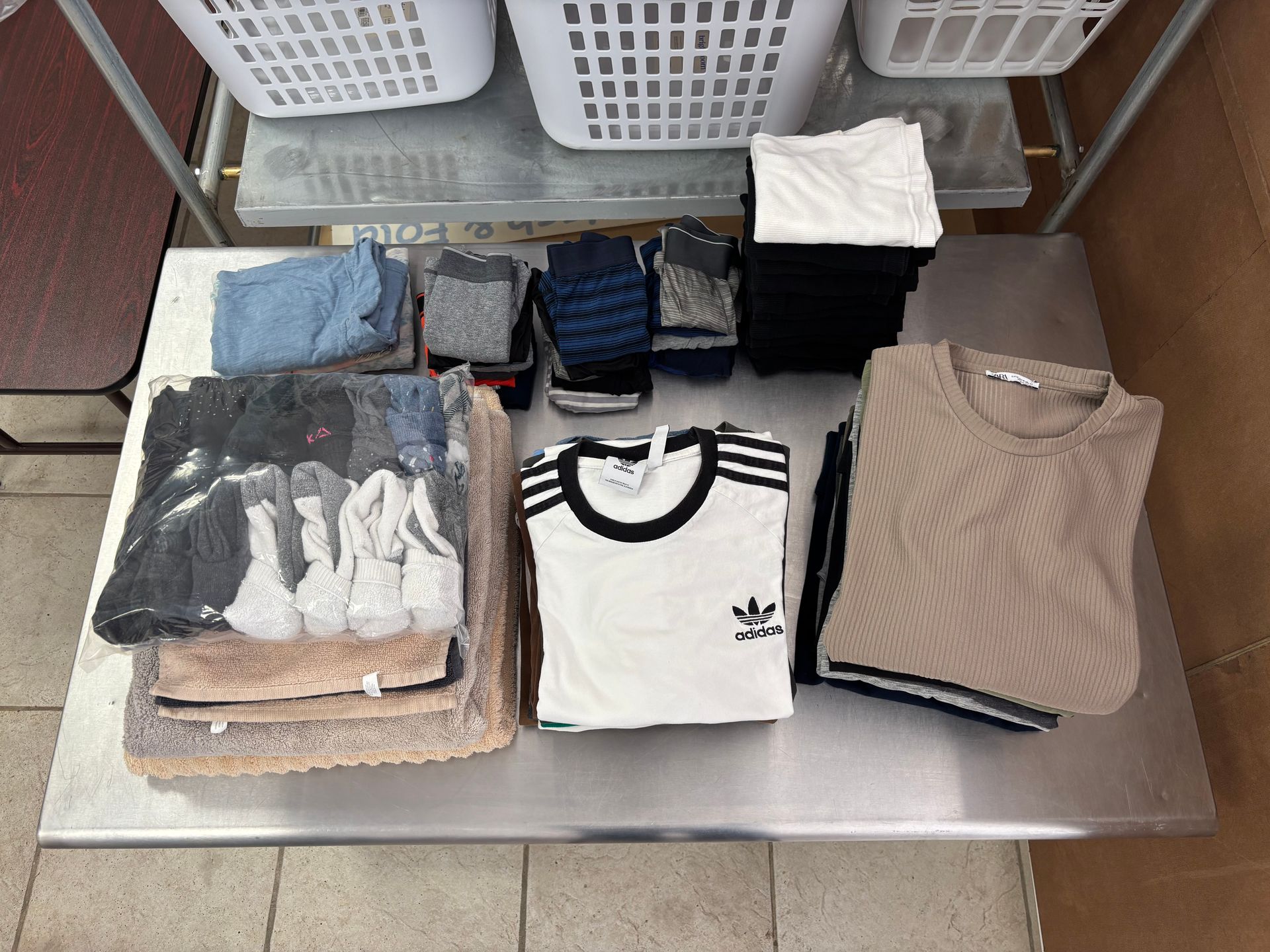 Folded laundry on a metal surface, including shirts, socks, and towels, with laundry baskets in the background.