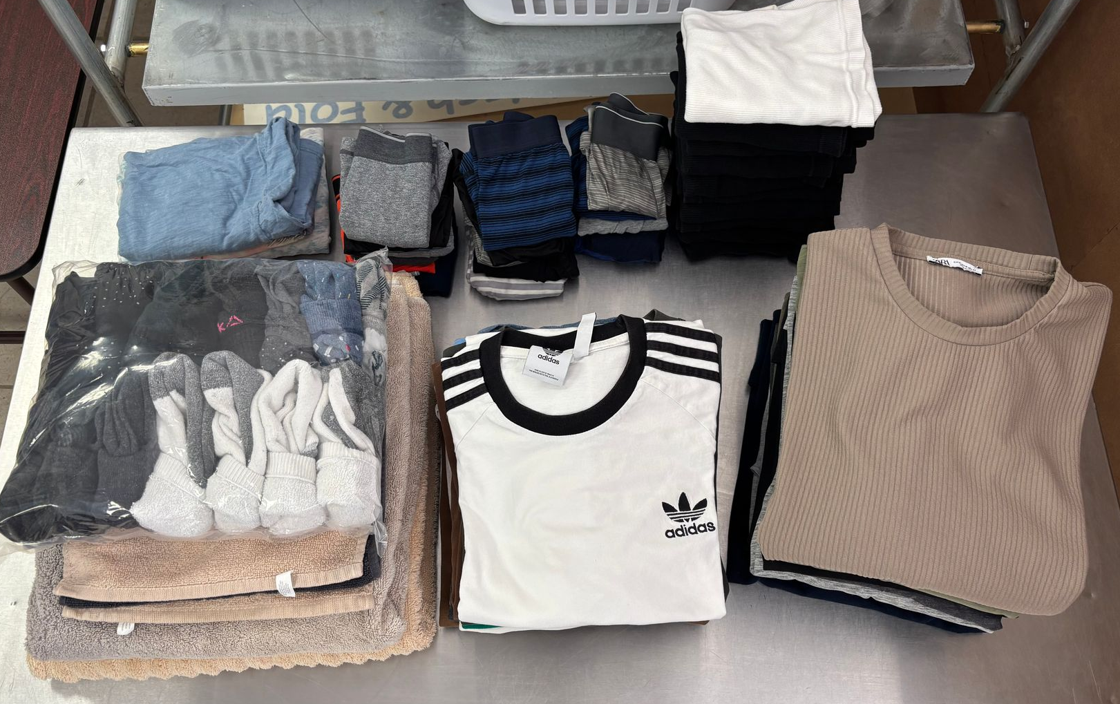 Folded laundry on a metal surface, including shirts, socks, and towels, with laundry baskets in the background.