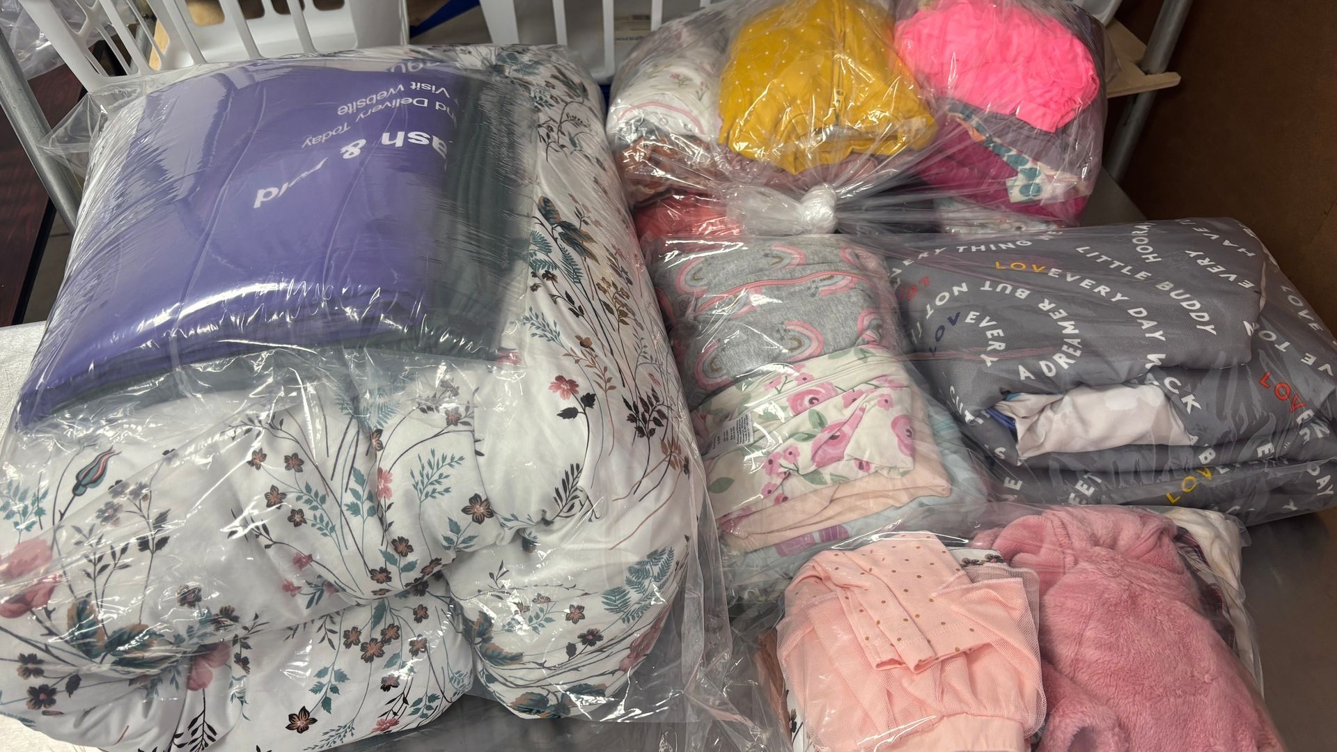 Assorted bedding sets, pillows, and blankets in clear plastic packaging on a table.