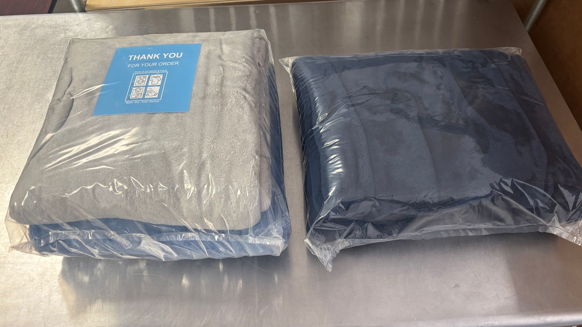 Two folded blankets, one gray and one blue, wrapped in plastic on a silver surface.