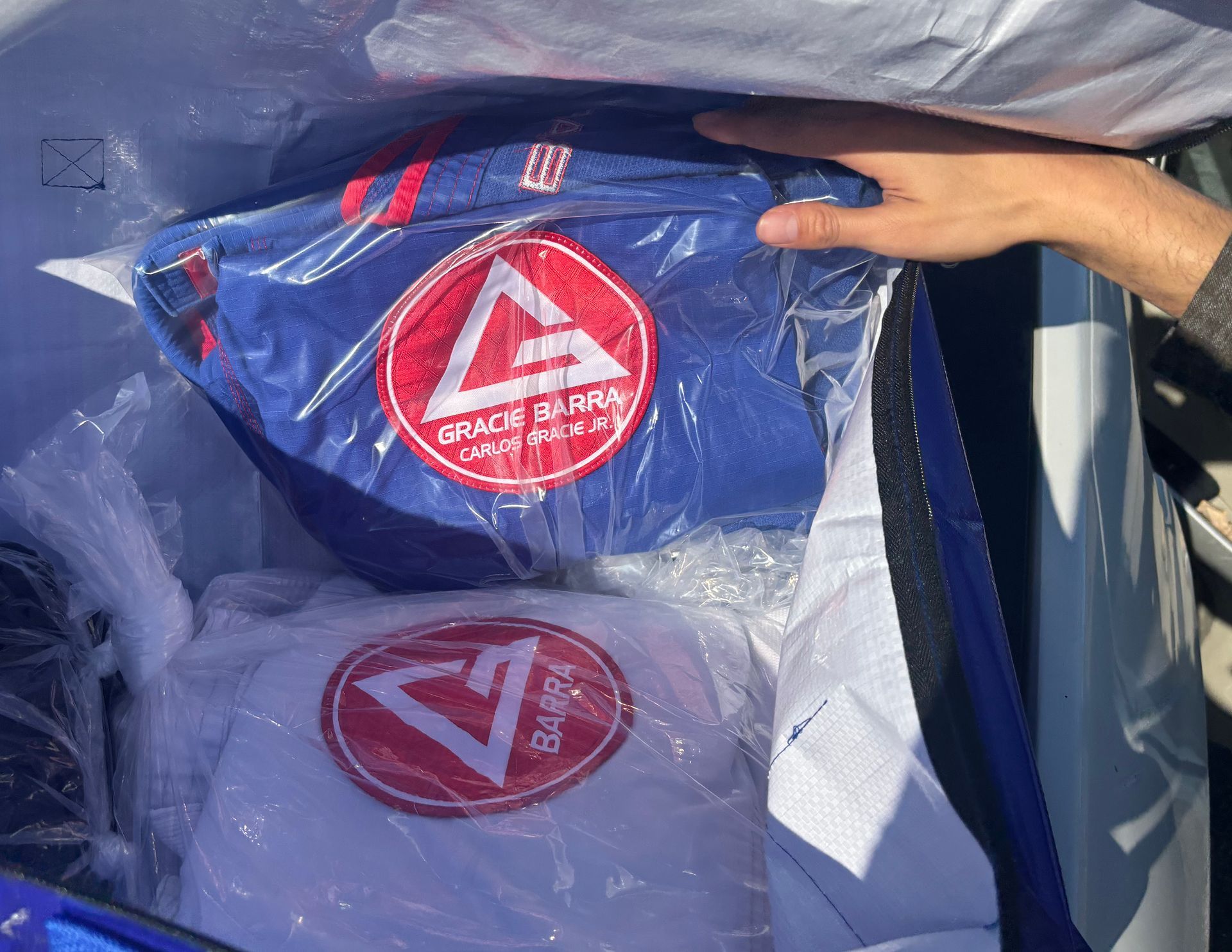Hand reaching into a bag containing a blue and white Gracie Barra Jiu-Jitsu gi, in plastic packaging.