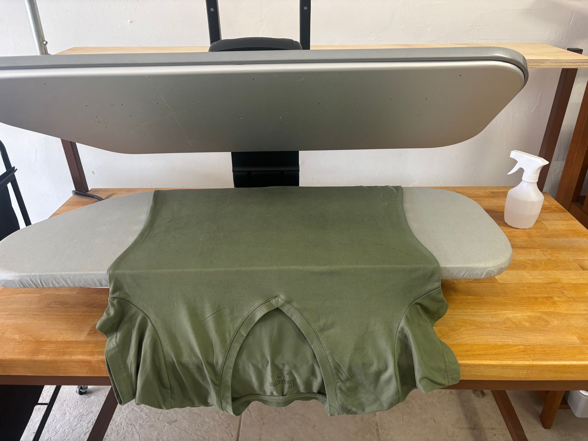 A green shirt being pressed on an industrial ironing machine on a wooden table; a spray bottle is nearby.