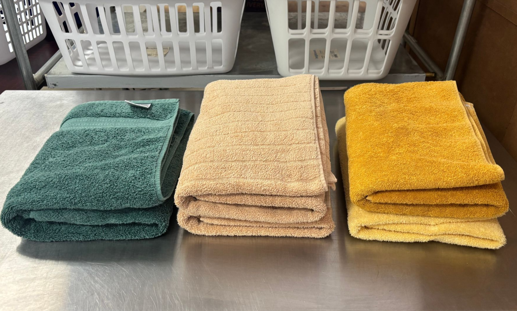 Three stacks of folded towels in green, tan, and yellow on a metal surface, with laundry baskets in the background.
