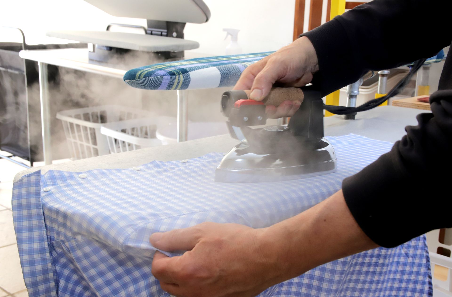 Person ironing a blue and white checkered shirt with a steam iron on an ironing board.