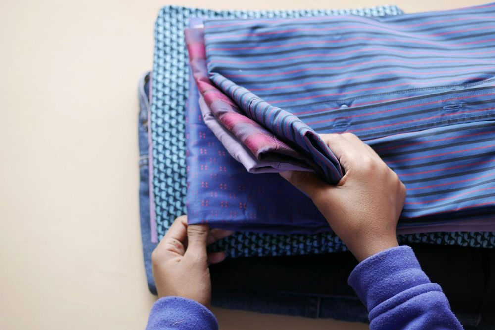 Hands folding a stack of colorful, neatly folded shirts on a light-colored surface.