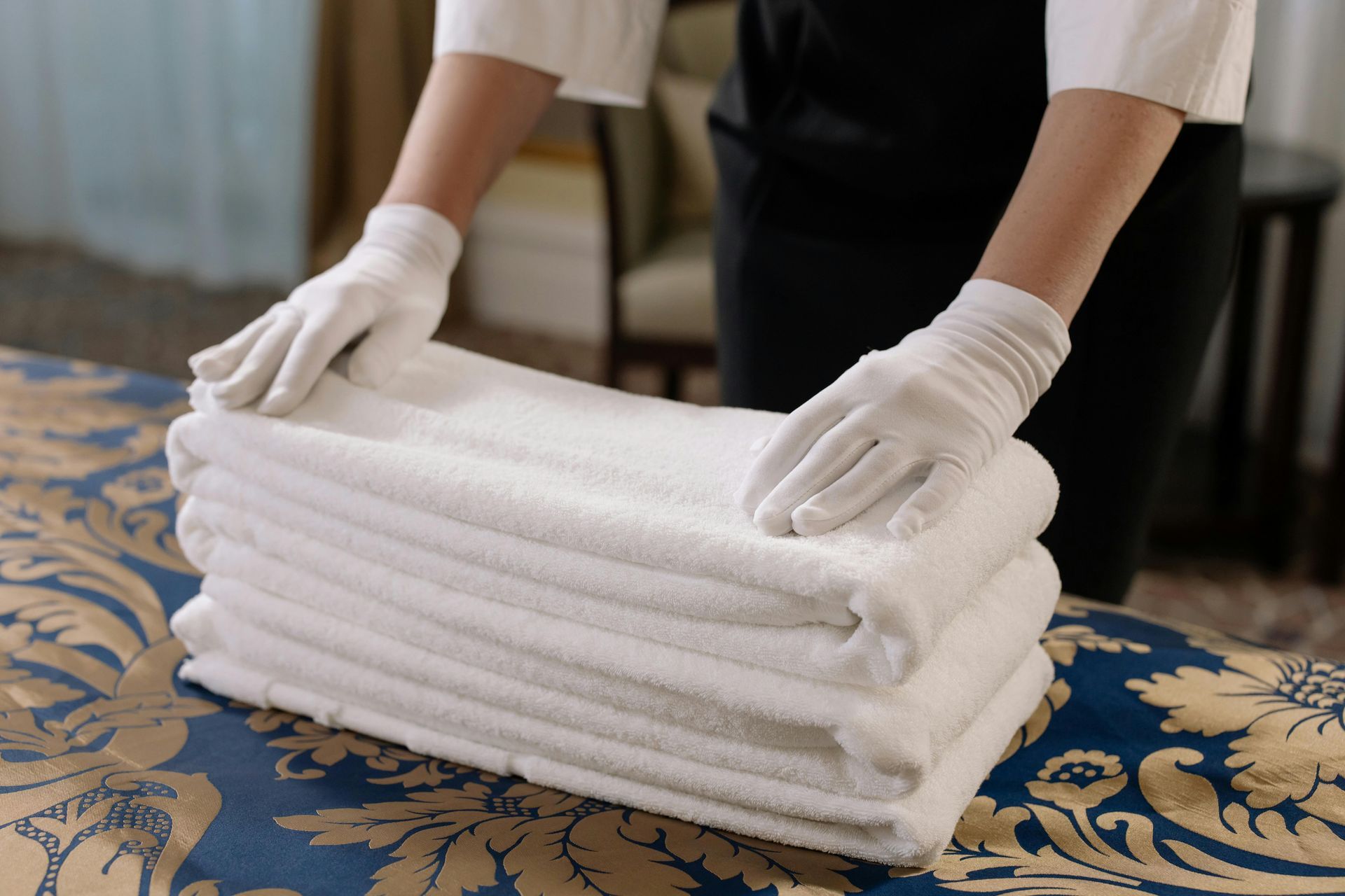 Person in white gloves folding white towels on a bed in a hotel room.