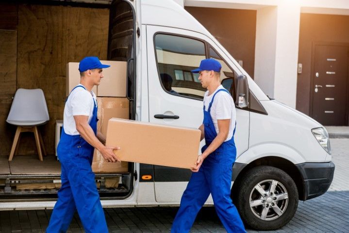 Two movers in blue overalls load a box into a white moving truck. Sunlight illuminates the scene.