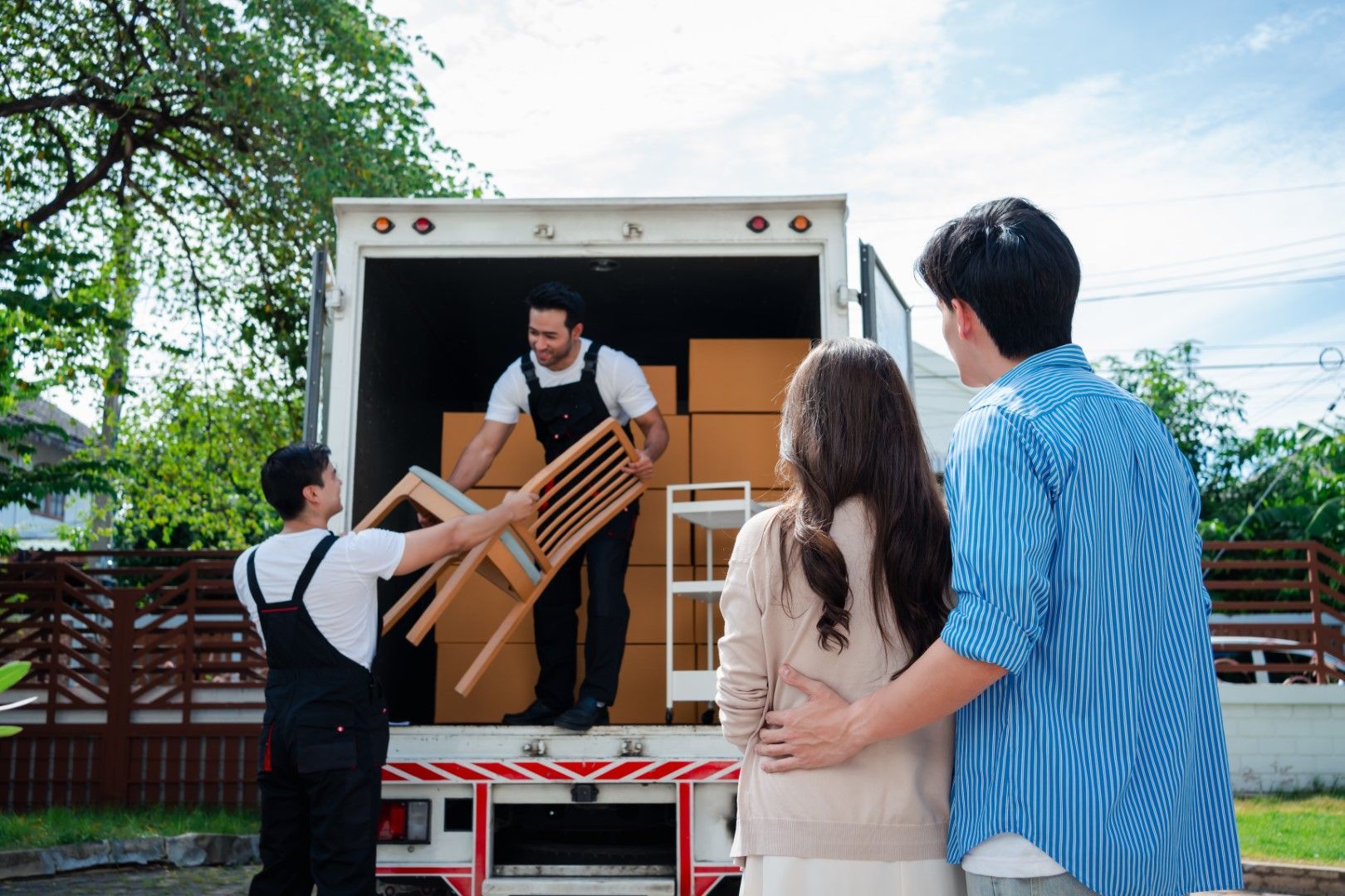 Movers loading a moving truck with furniture and boxes, while a couple watches in front of a house.
