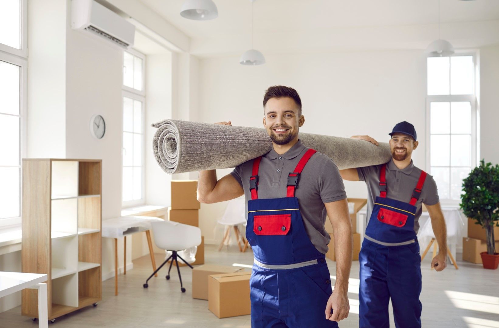 Two movers wearing blue overalls carry a rolled-up rug through a bright, empty room. They smile at the camera.