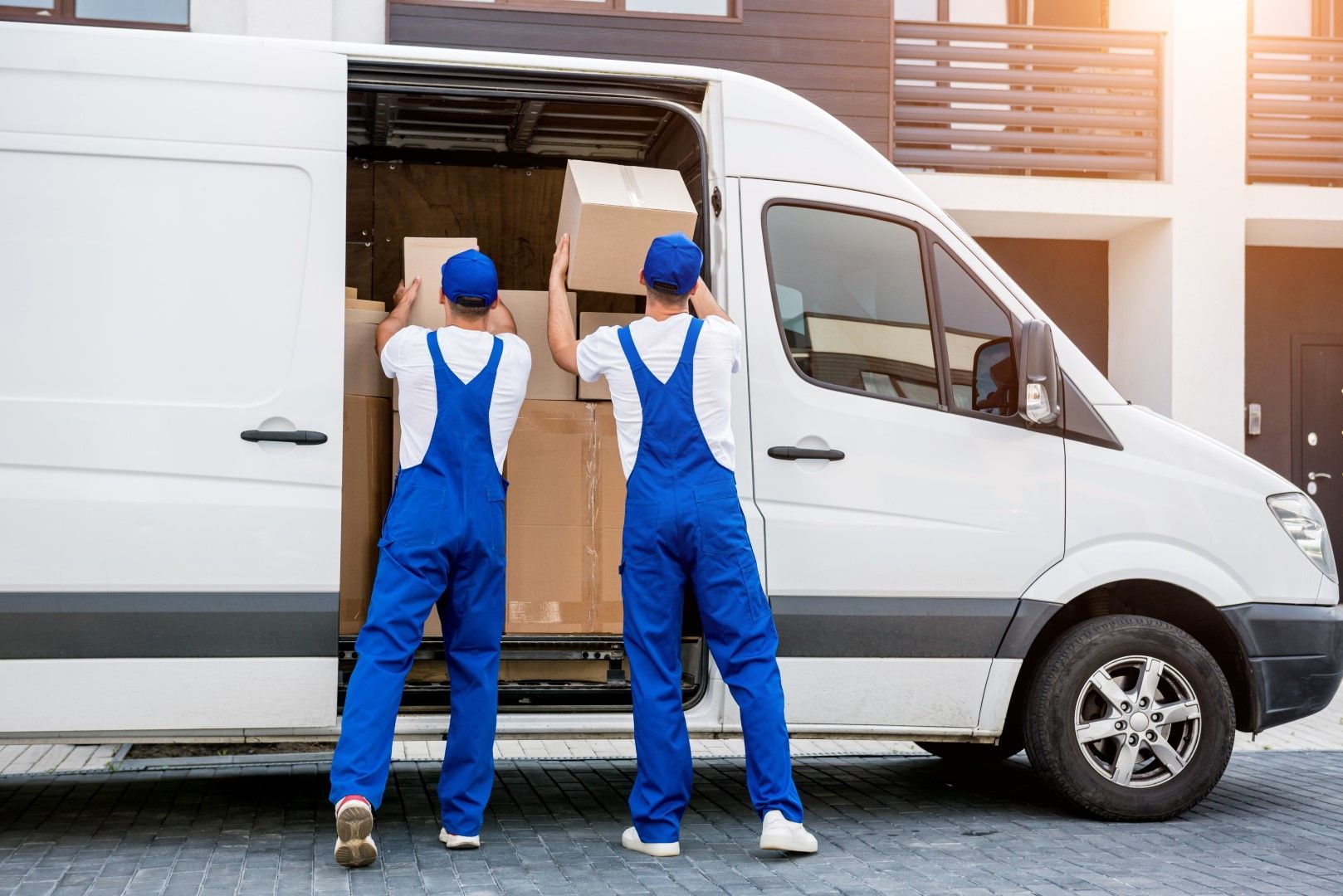 Two movers in blue coveralls load cardboard boxes into a white delivery van.