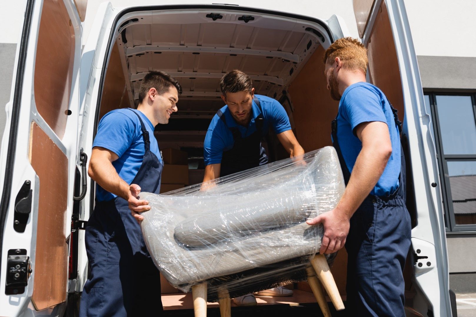 Three movers in blue shirts loading a wrapped chair into a white van. The movers are focused and working together.