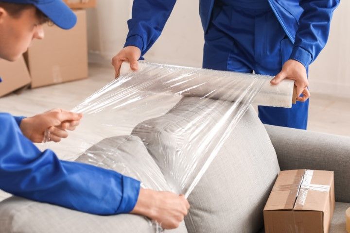 Two movers in blue uniforms wrapping a gray sofa in plastic wrap, indoors. One person pulls the wrap taut while the other holds the roll.