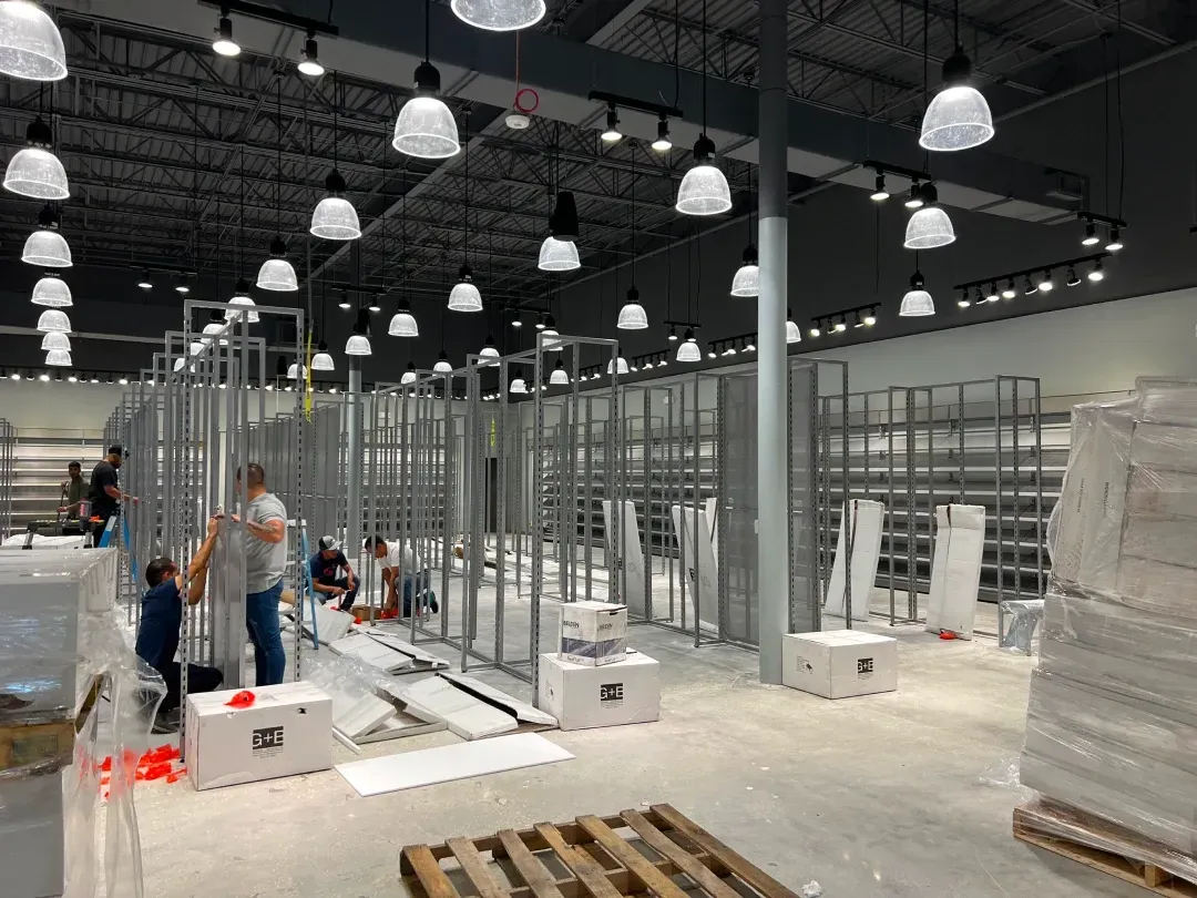 Construction workers installing shelving inside a large retail space. Rows of metal frames are in place, with overhead lighting.