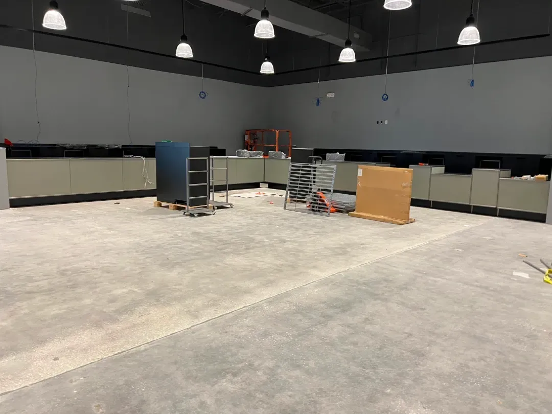 Empty commercial space with gray walls, concrete floor, and overhead lights; cabinets line walls, construction materials present.