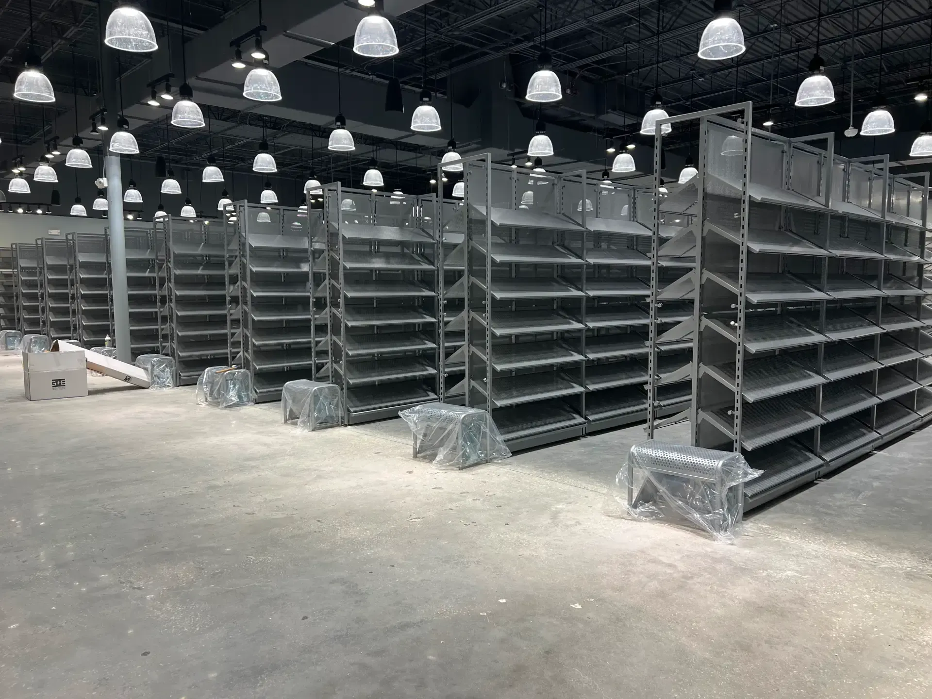 Rows of empty gray retail shelving units in a large warehouse-like space, lit by overhead lights.