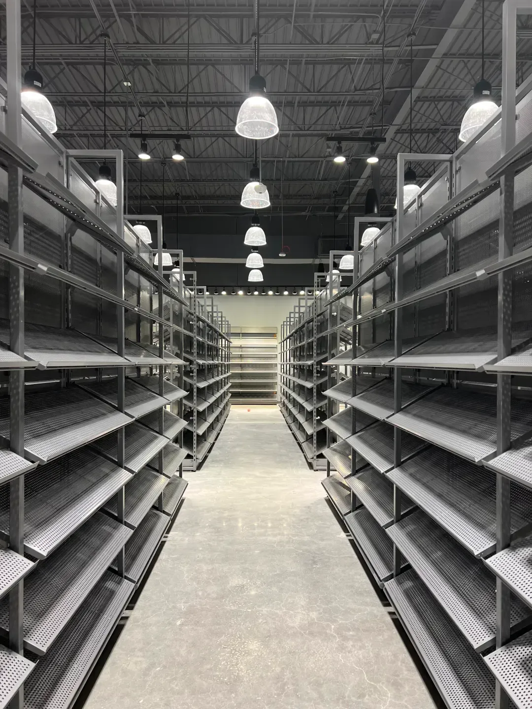 Empty gray shelves in a warehouse setting, receding into the distance. Overhead lights.