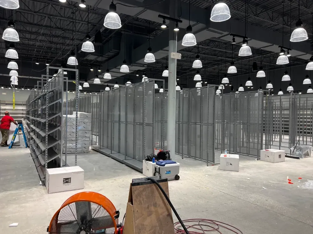 Interior view of a large warehouse-like space with metal shelving units and overhead lighting. A worker is present.