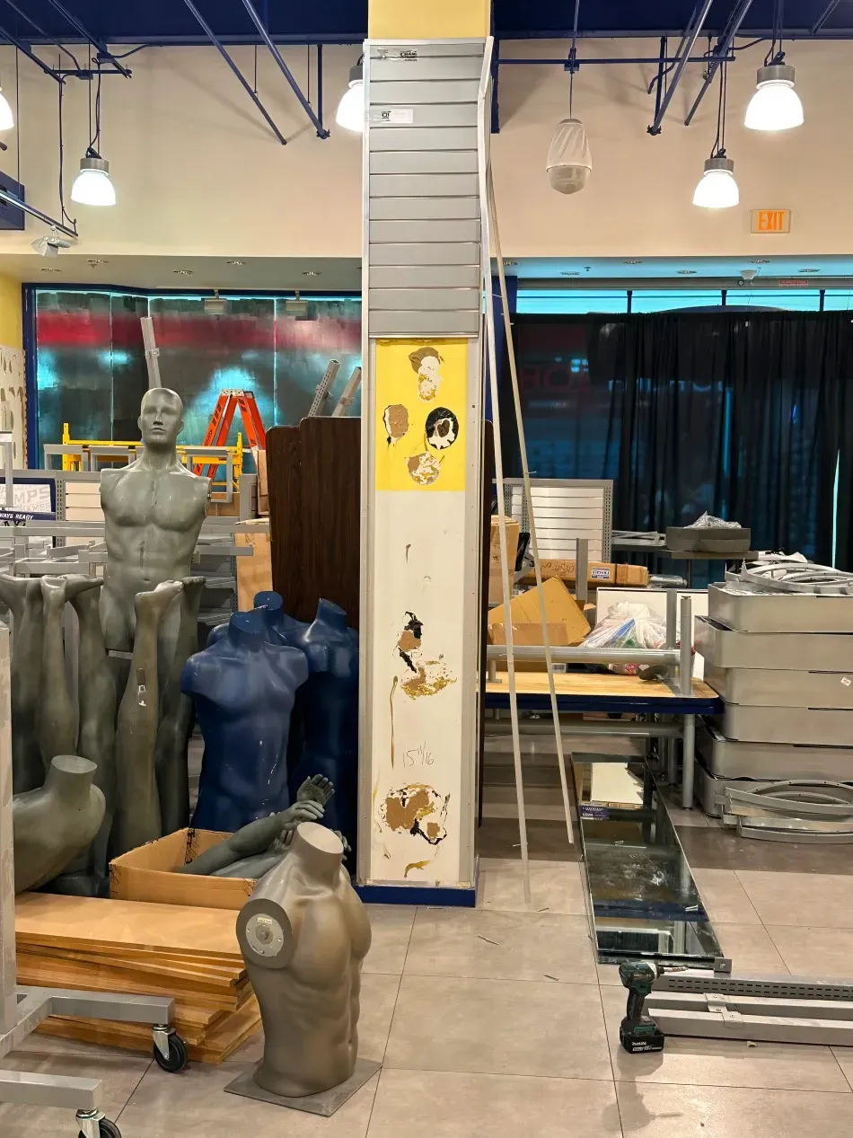 Interior view of a store with display mannequins and sculptures. A central support column features peeling paint and a cartoon.