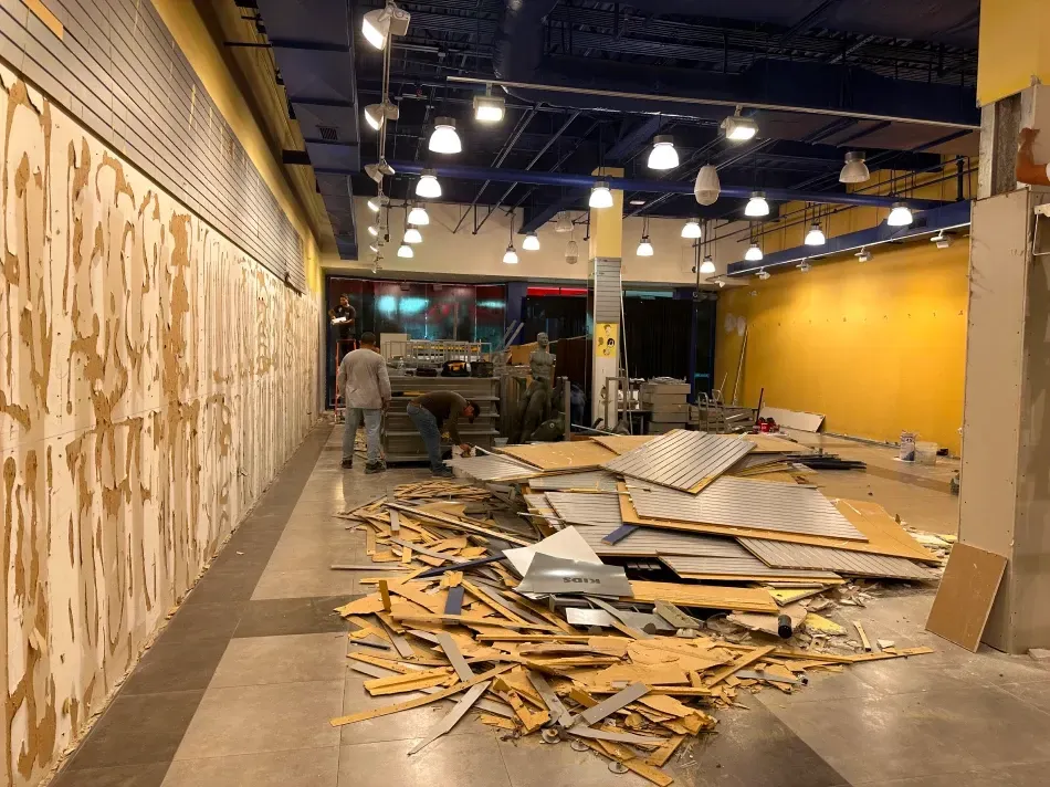Interior demolition: debris pile in foreground, workers, yellow and blue walls, overhead lights.