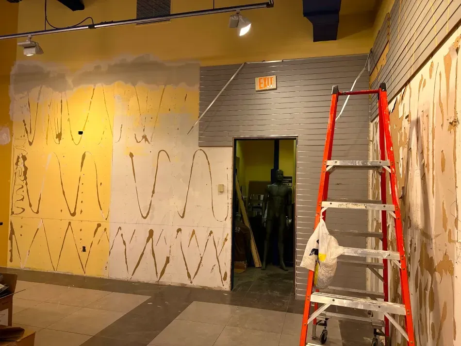 Interior view of a room under renovation. A ladder stands by a wall with exposed surfaces and an exit sign above a doorway.
