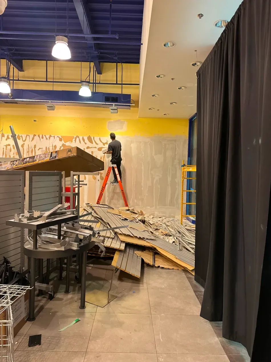 Man on a ladder removing a wall in a retail space under renovation. Debris and yellow wall visible.