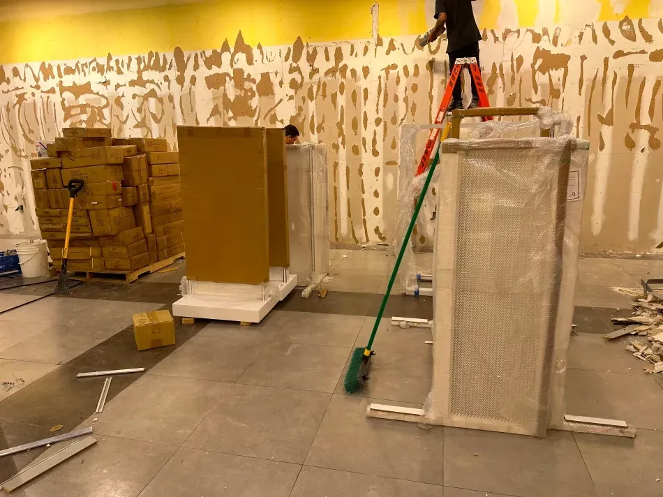 Construction scene: Unboxed door frames, stacked boxes, worker on a ladder, debris on tiled floor, and stripped wallpaper.