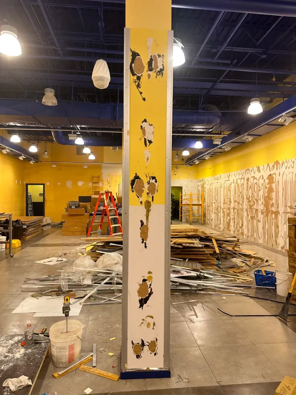 Interior of a retail space undergoing renovation. Walls are partially torn down, debris on the floor, and a central yellow column with paint damage.