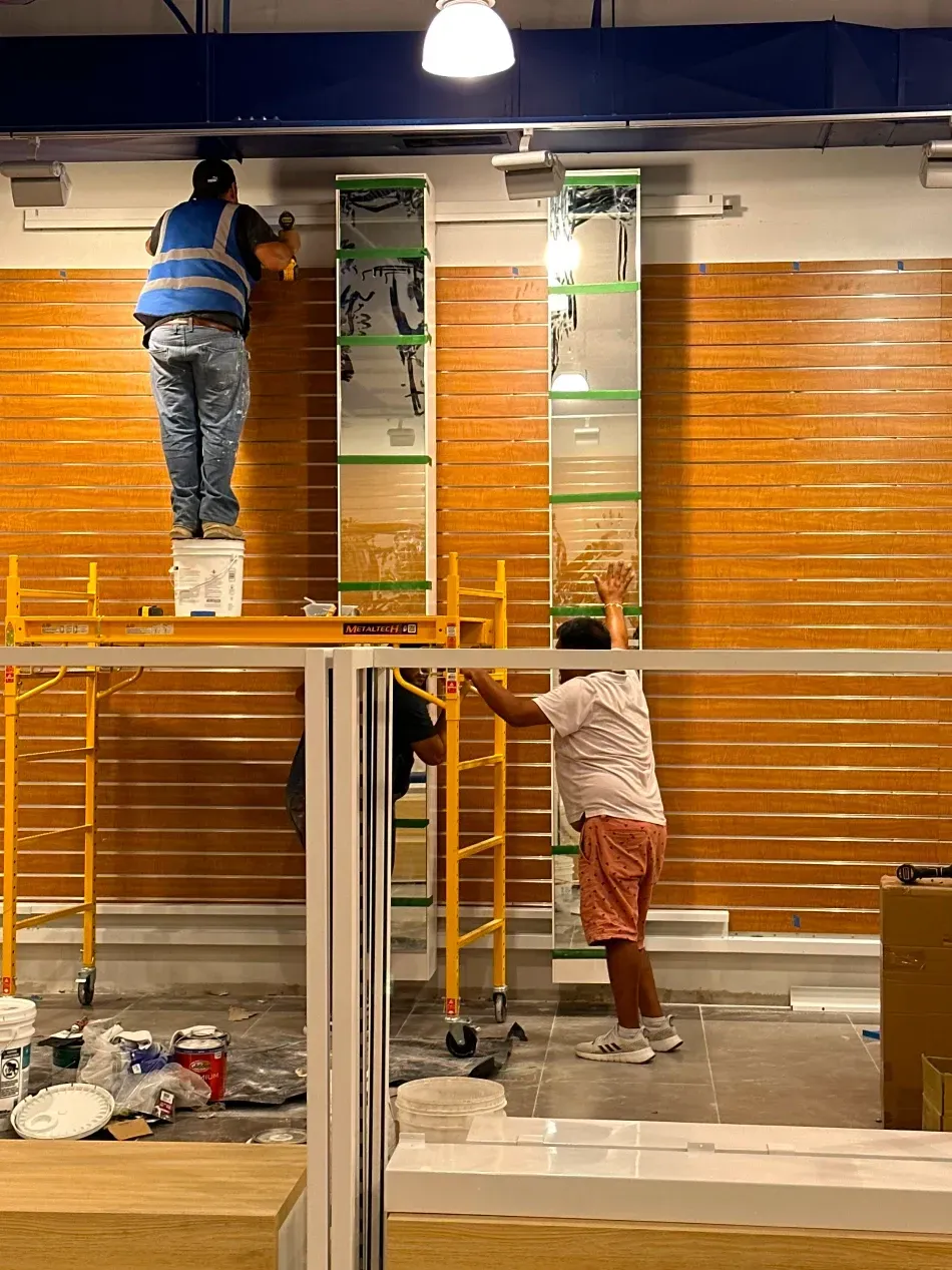 Two people painting a wooden wall, using a scaffold. Inside a building under construction.