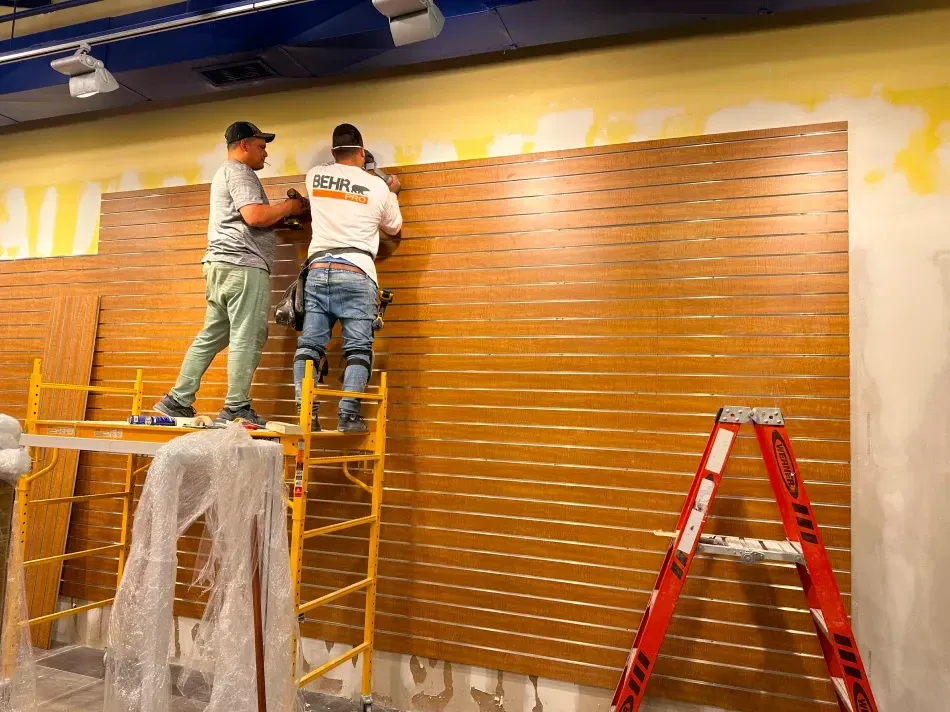 Two workers installing wood paneling on a wall, using scaffolding and a ladder in a room.