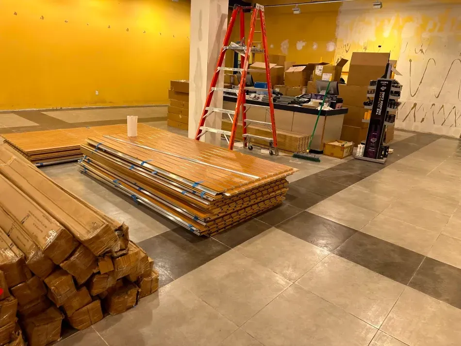 Construction site: stacked wood flooring and boxes on tiled floor. Orange ladder near yellow wall.