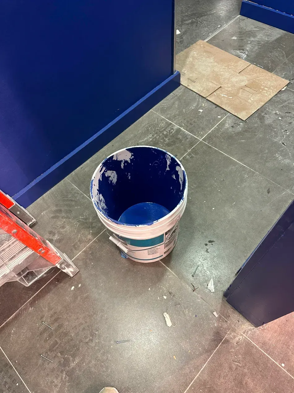 Paint bucket of blue paint next to a partially painted blue wall and small cardboard sheet on floor.