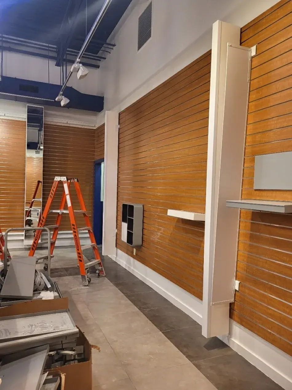 Interior retail space under construction, wood-slat walls, white support beam, ladder, and concrete floor.