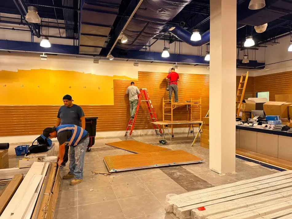 Construction workers installing wood paneling on a yellow wall inside a building.