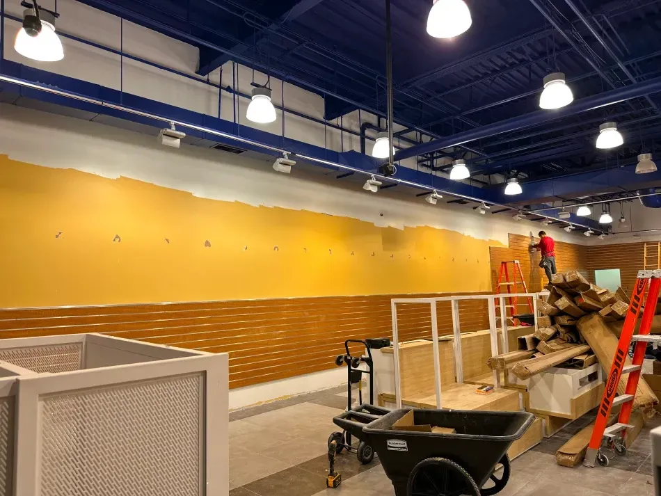 Interior of a store under construction; yellow walls, blue ceiling, construction materials, and a worker on a ladder.