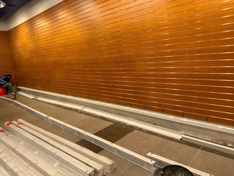 Construction in a room with a wood-slat wall. Workers assemble metal tracks on the floor.