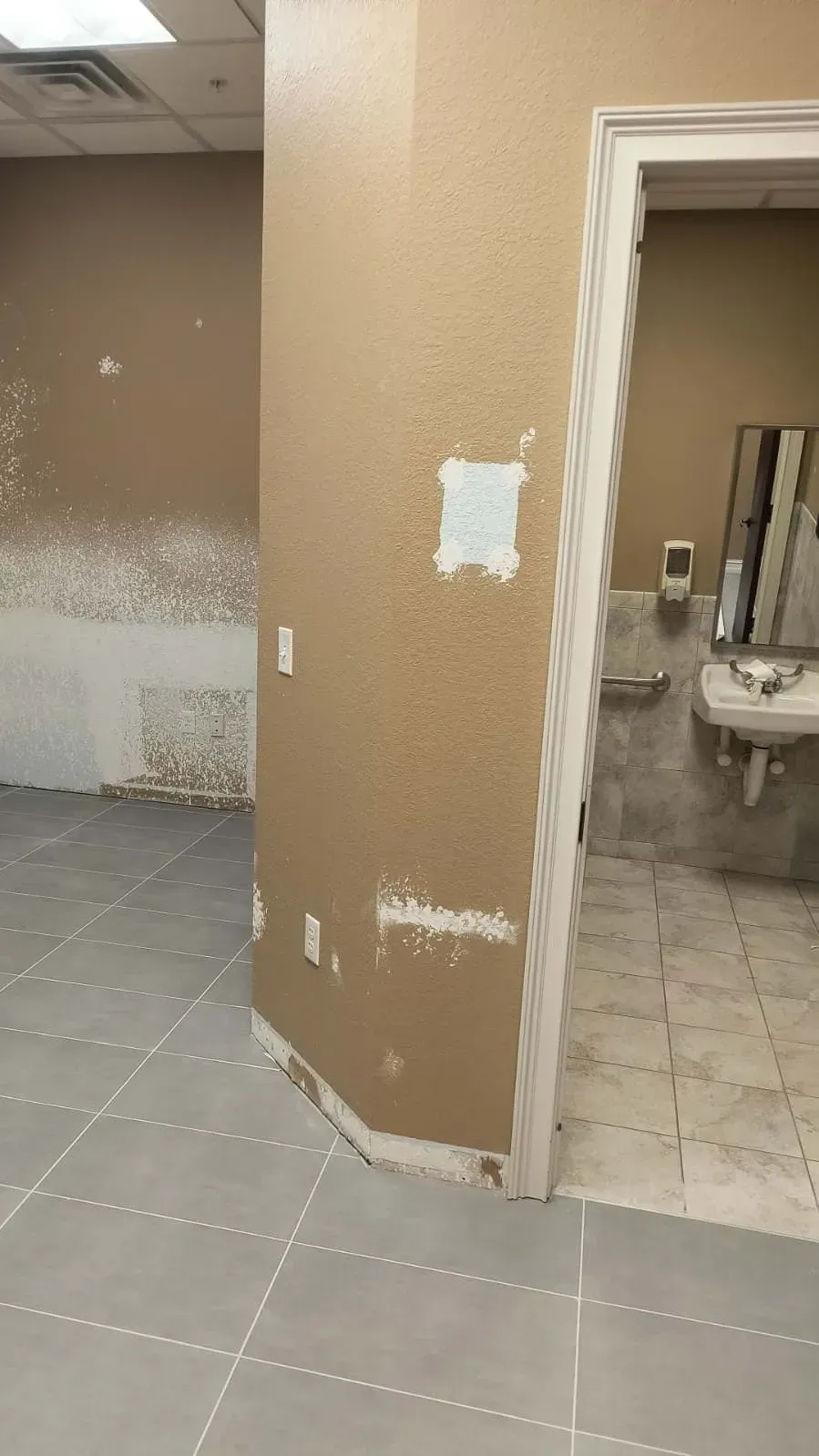 Interior view of a bathroom under renovation. Tan walls with peeled paint, gray tile floor, and doorway to a restroom with a sink.