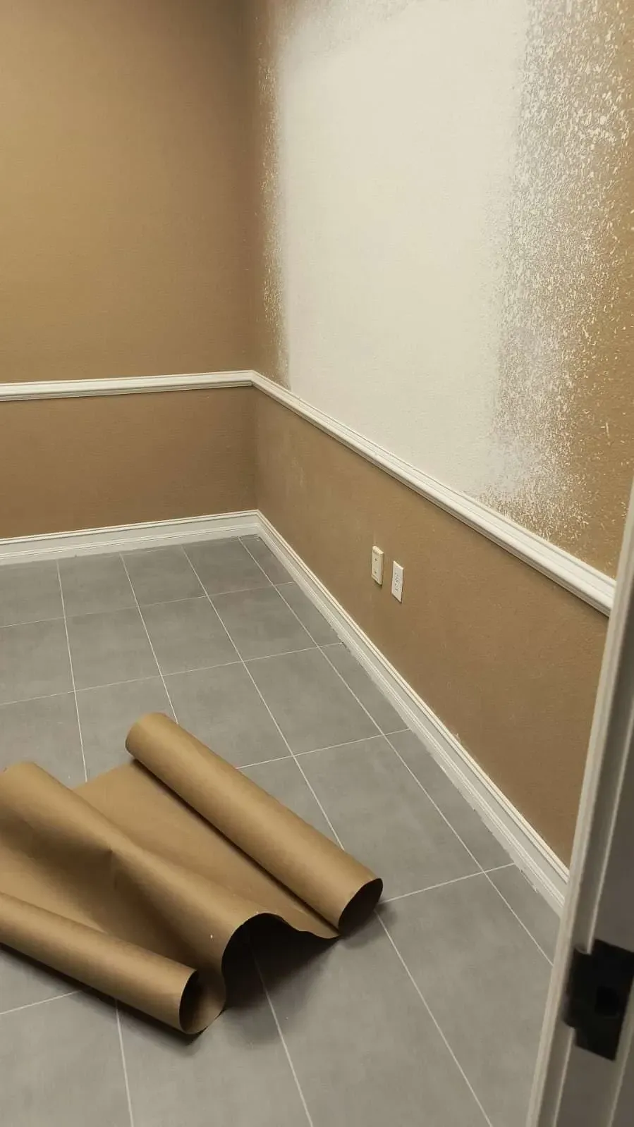 Room being painted; white paint on tan wall, gray tiled floor, rolled brown paper in foreground.