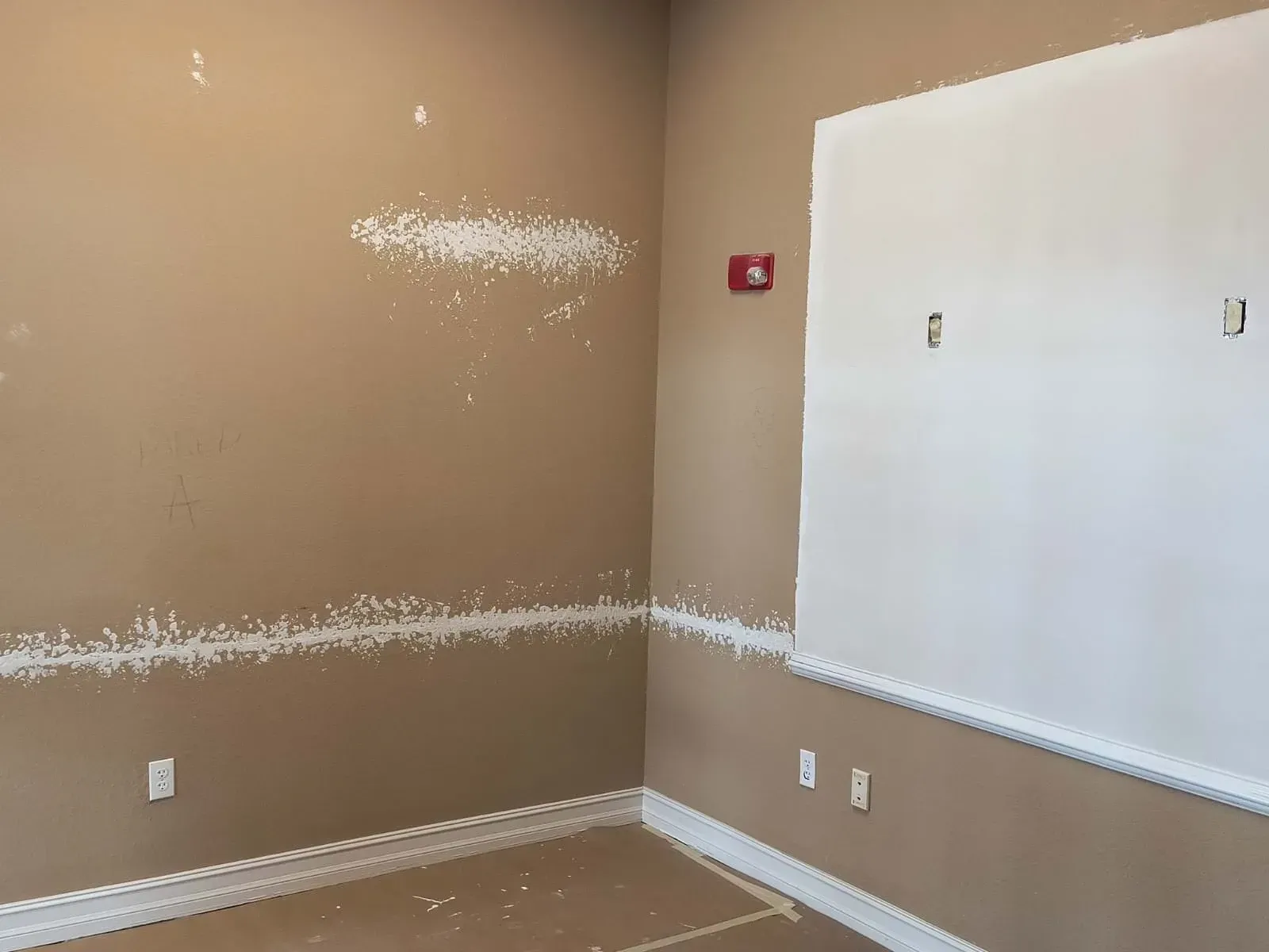 A room with tan walls, white baseboards, and a white-painted section near a window. Fire alarm on wall.