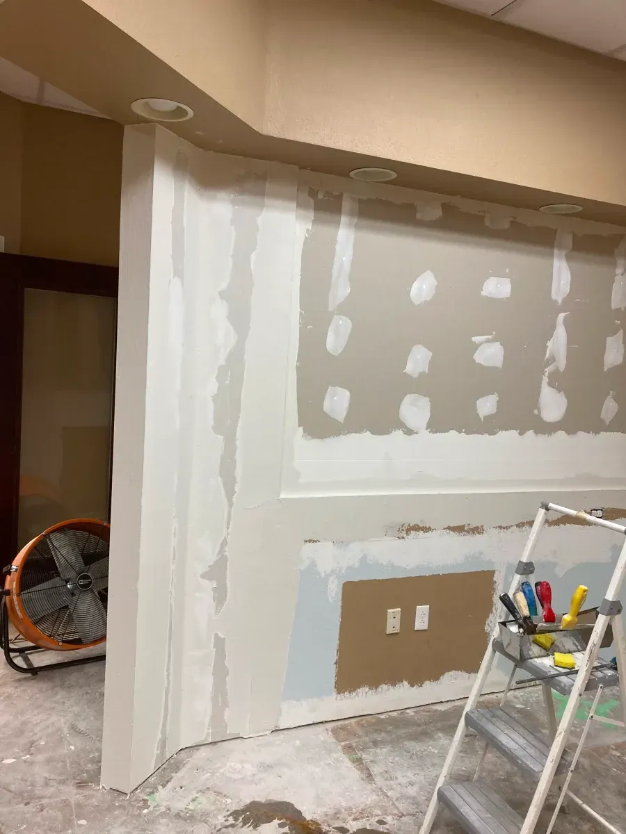 Interior wall under renovation, with drywall, mudding, and a stepladder. A fan is visible.