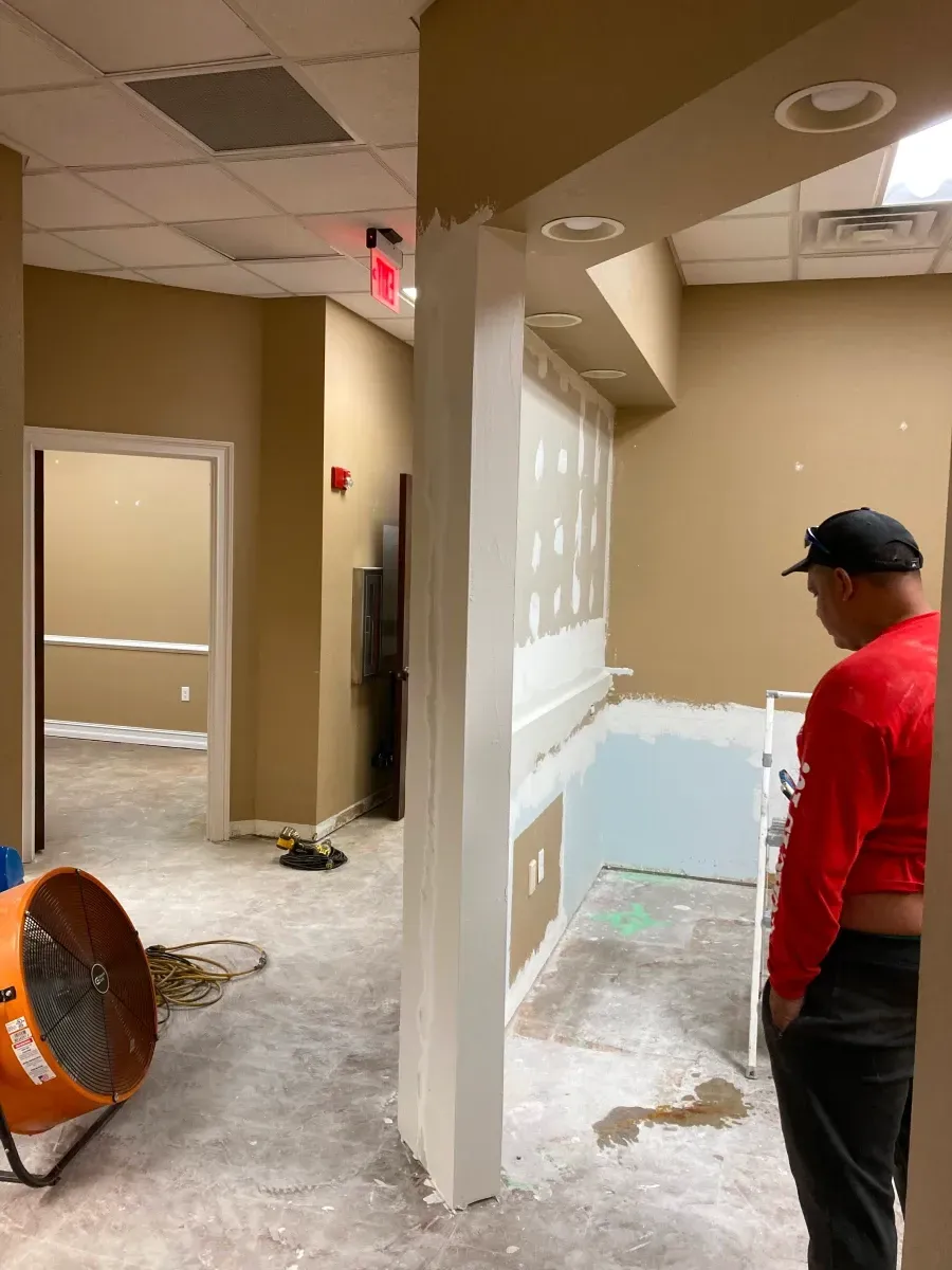 Person standing in a room under renovation, looking at a wall. An orange fan is on the floor.