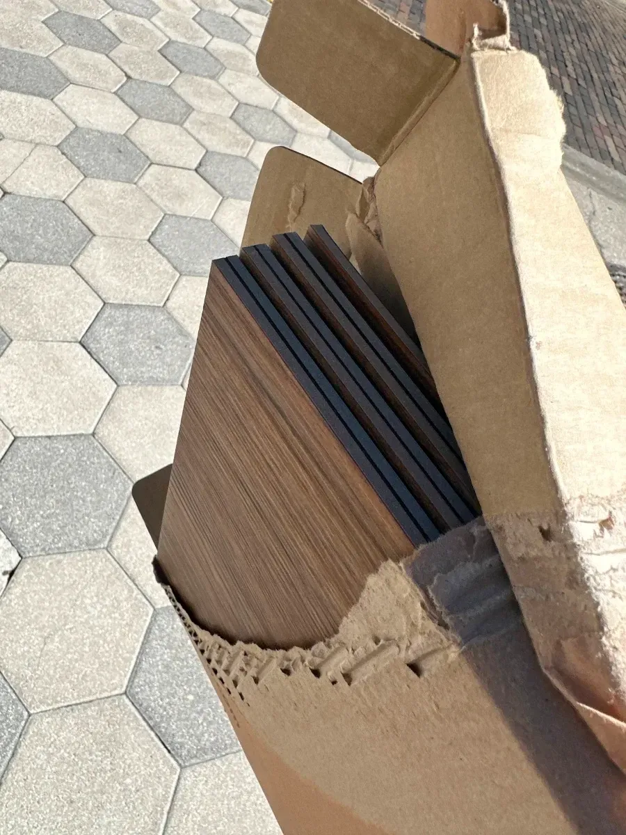 Cardboard box open, containing stacked brown wood-look planks, on a patterned hexagonal stone surface.
