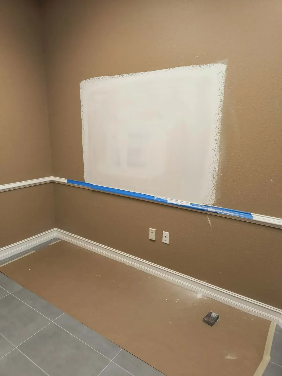 A room corner with brown walls, white patch, blue tape, and gray floor tiles.