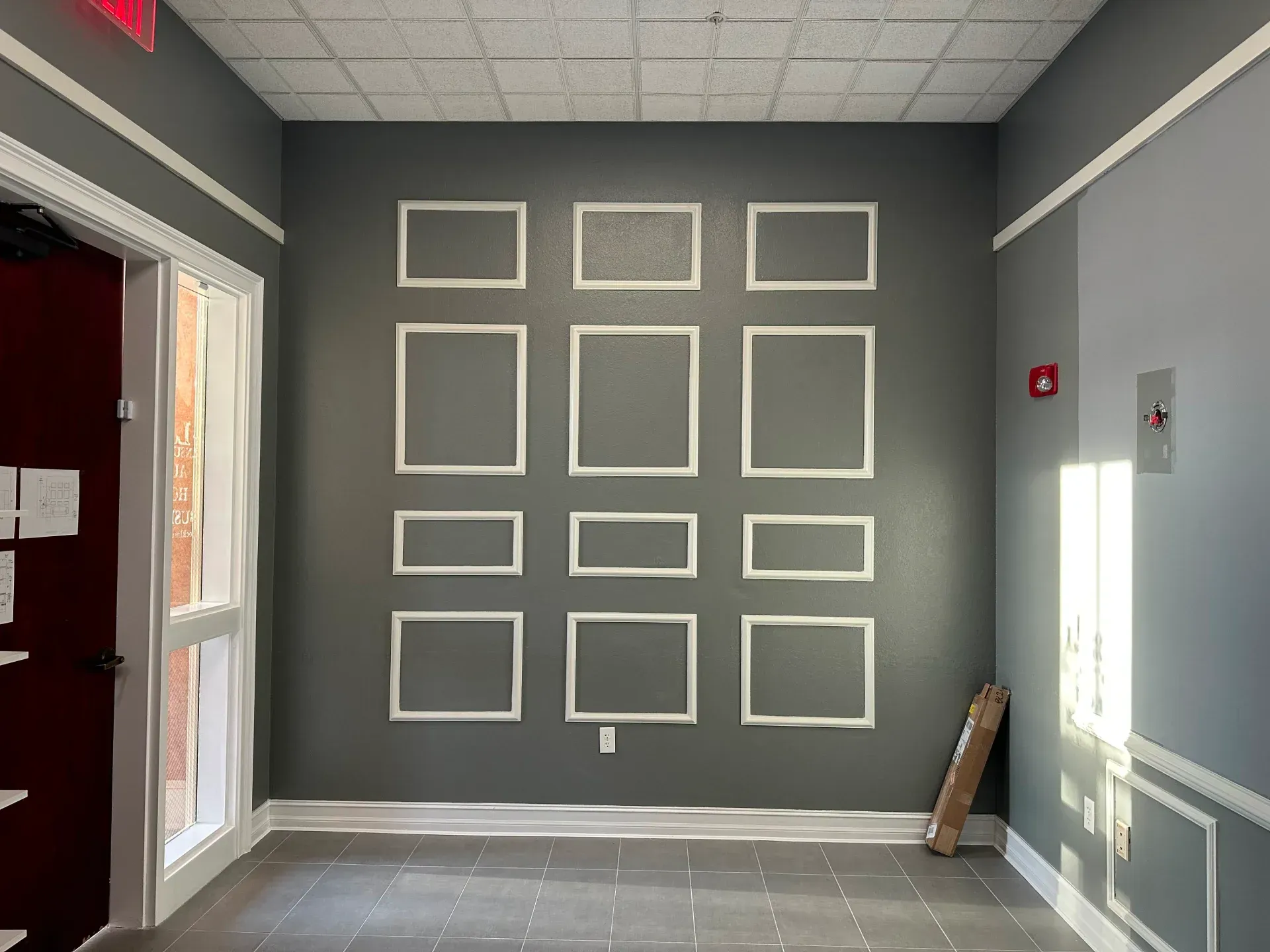 Gray-walled interior with decorative white frames. A doorway and window are on the left; an elevator and light on the right.