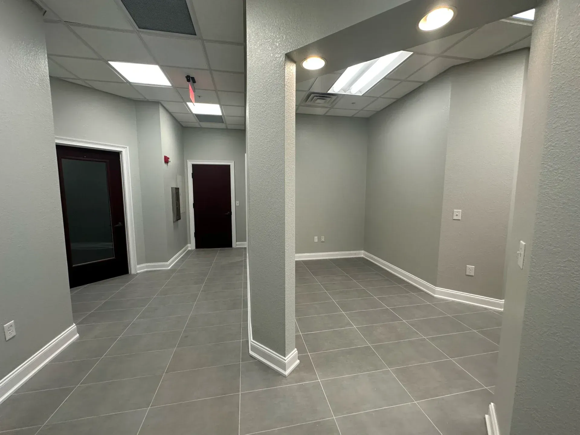 Empty office space with gray walls, tile floors, and a column. Dark doors at the end of the hallway.