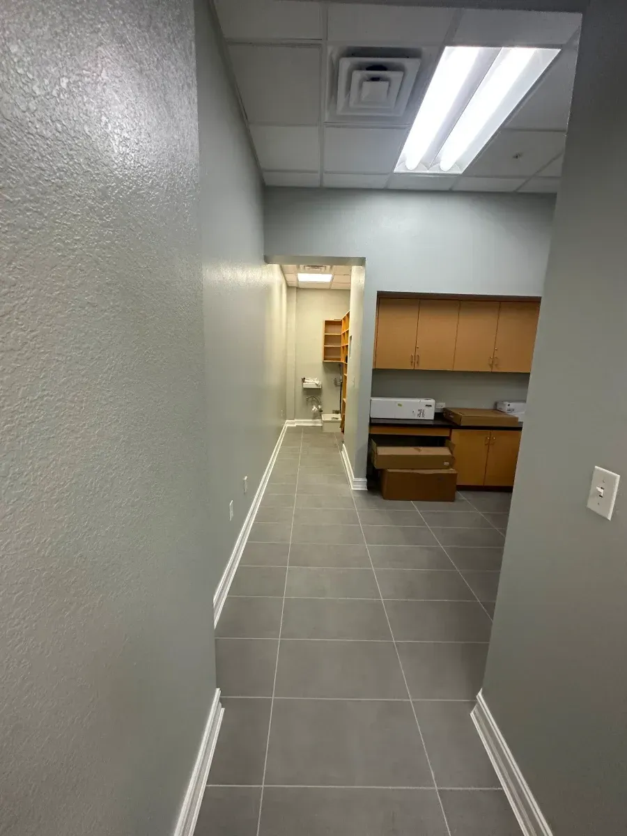 Narrow office hallway with gray walls, tile floor, and a doorway leading to a utility space.