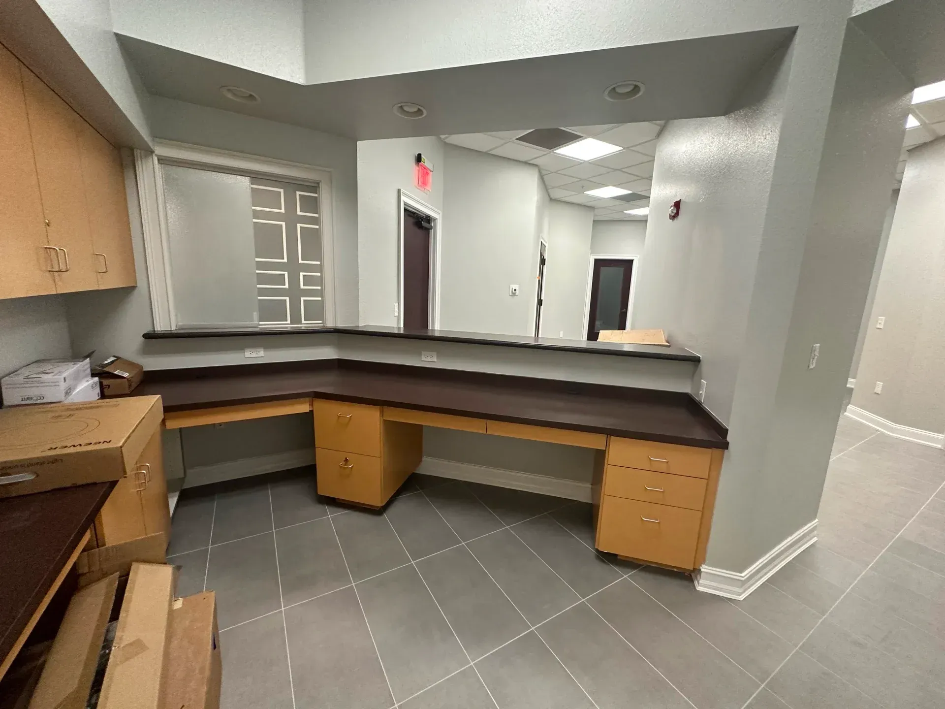Reception desk in an office. Wooden cabinets, dark countertop, gray tile floor. Boxes are on the left.