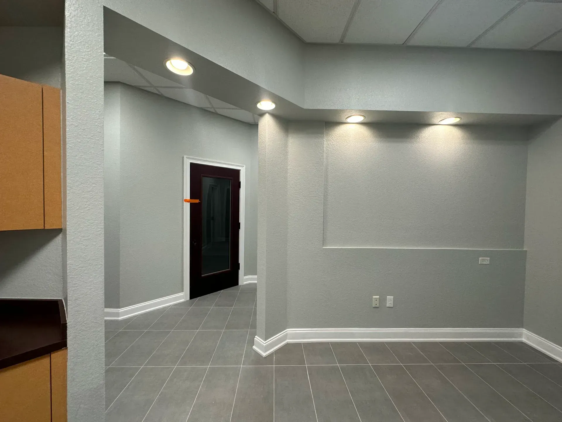 Empty room with gray walls, recessed lighting, and a dark door in the background. Brown cabinets on the left.