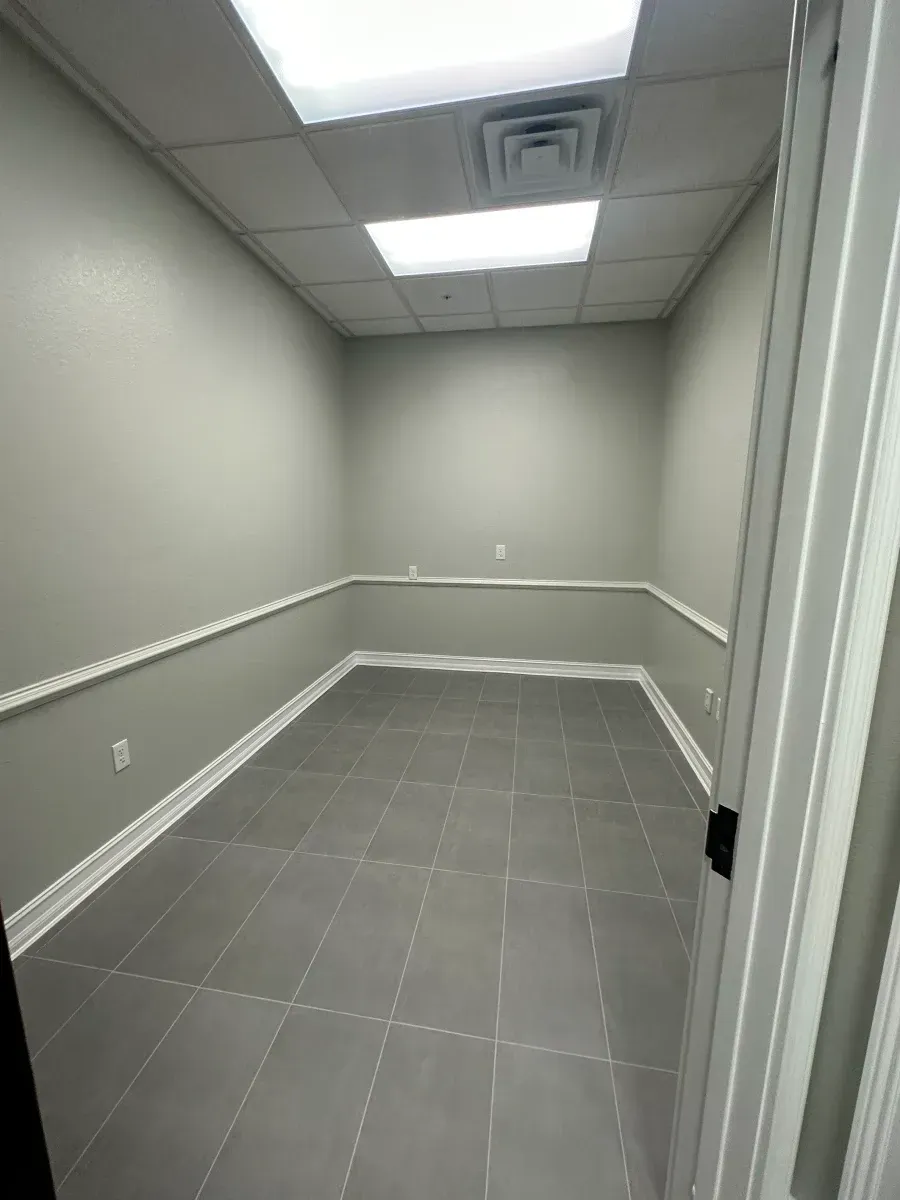 Empty office room with gray walls, tile floor, and white trim. Bright overhead lighting.