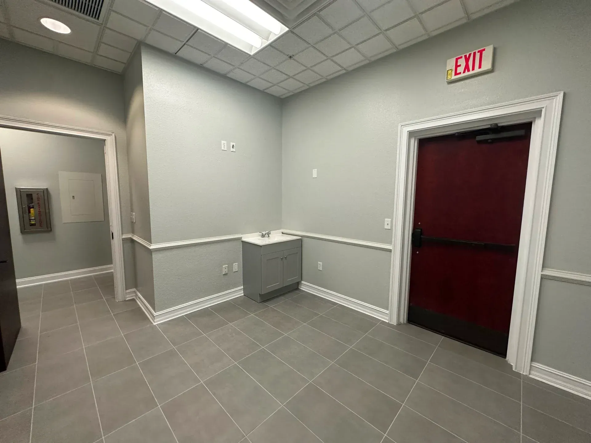 Empty office interior with gray tile floor, gray walls, red door and exit sign.